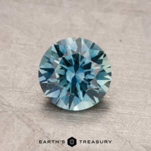 1.14-carat round brilliant-cut teal Montana sapphire on a textured surface with Earth's Treasury logo at the bottom.