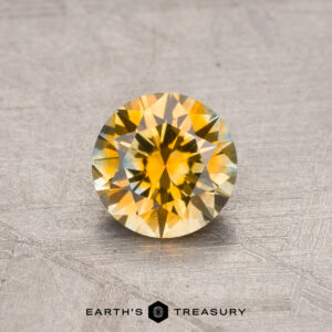 A round yellow Montana sapphire with a faceted cut, weighing 0.86 carats and heat-treated, is displayed on a textured gray surface. The text "Earth's Treasury" appears at the bottom of the image.