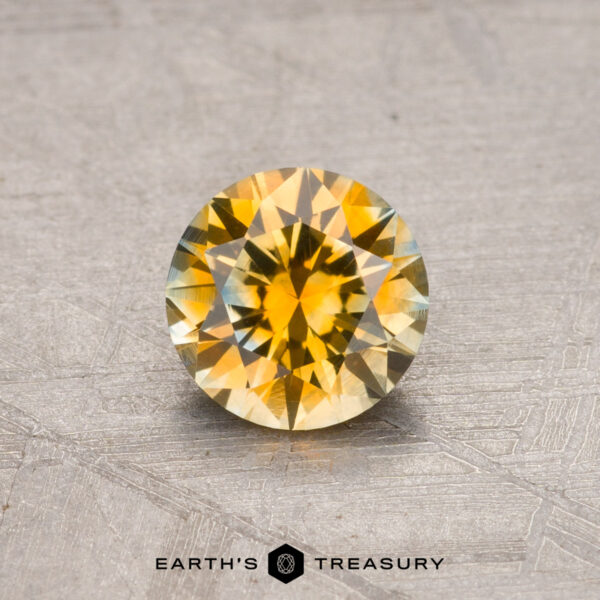 A round yellow Montana sapphire with a faceted cut, weighing 0.86 carats and heat-treated, is displayed on a textured gray surface. The text "Earth's Treasury" appears at the bottom of the image.