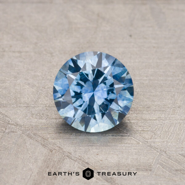 A round blue 0.93-carat Montana sapphire with a faceted cut is placed on a textured surface, accompanied by the text "Earth's Treasury" at the bottom.