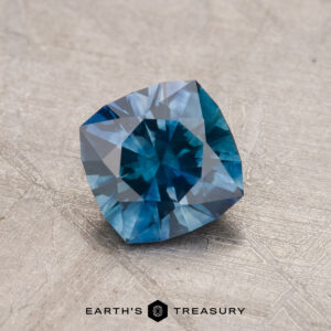 A close-up of a cushion-cut blue Montana sapphire, weighing 1.03 carats and heated to enhance its beauty, showcasing its facets on a textured gray surface with the "Earth’s Treasury" logo at the bottom.