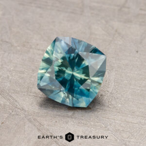 A square-cut, teal 1.02-Carat Montana Sapphire (Heated) rests on a textured surface, with the "Earth's Treasury" logo at the bottom.