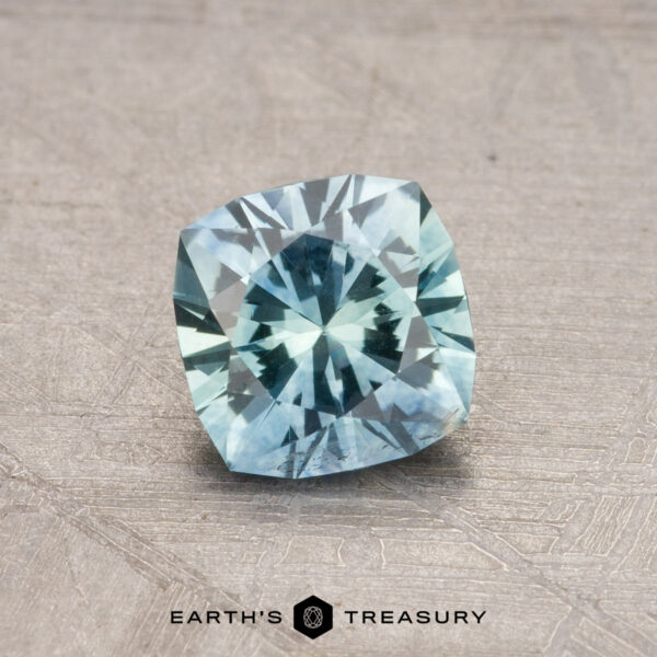 A 0.92-carat Montana Sapphire (heated) with a pale blue hue shines while resting on a textured surface, accompanied by the "Earth's Treasury" logo beneath it.