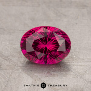 A vibrant, oval-cut pink gemstone with intricate facets rests on a textured surface, marked with "1.63-Carat Ceylon Ruby (Heated)" at the bottom.