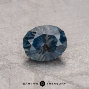 A faceted round teal 1.01-carat Montana sapphire gemstone on a textured gray surface with the logo "Earth’s Treasury" beneath.