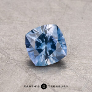 Displayed on a textured gray surface with the Earth's Treasury logo is a 0.91-carat, cushion-cut Montana sapphire gemstone.