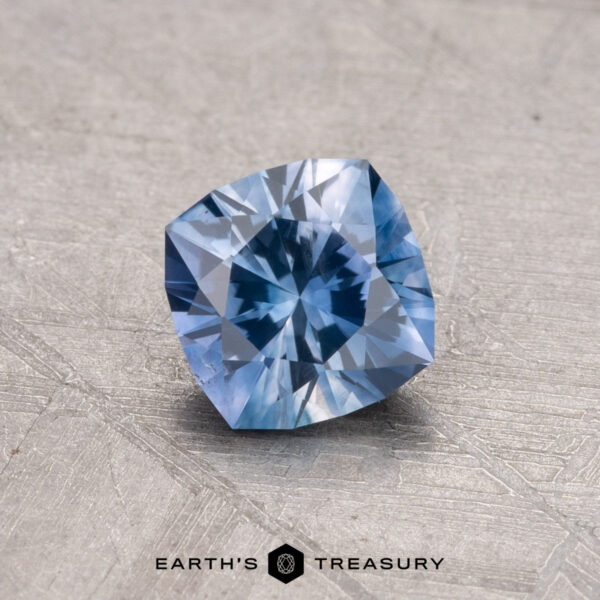 Displayed on a textured gray surface with the Earth's Treasury logo is a 0.91-carat, cushion-cut Montana sapphire gemstone.