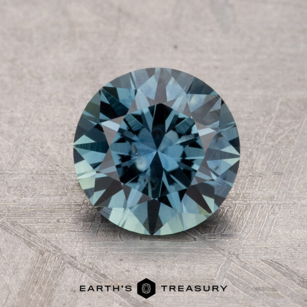 A round, faceted 1.53-carat Montana sapphire gemstone on a textured gray surface, branded with "Earth's Treasury" at the bottom.