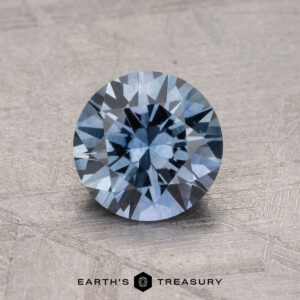 A round, faceted blue gemstone is placed on a textured surface with the text "1.36-Carat Montana Sapphire (Heated)" beneath it.